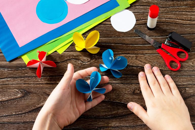 Close up of a child's hands on a table making paper butterflies with safety scissors and a stapler