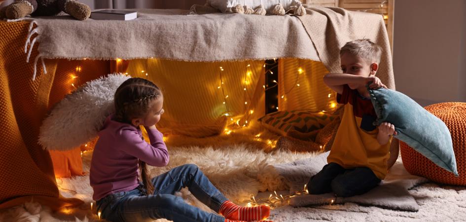 A young girl and boy sitting in front of a den with fairy lights draped over it