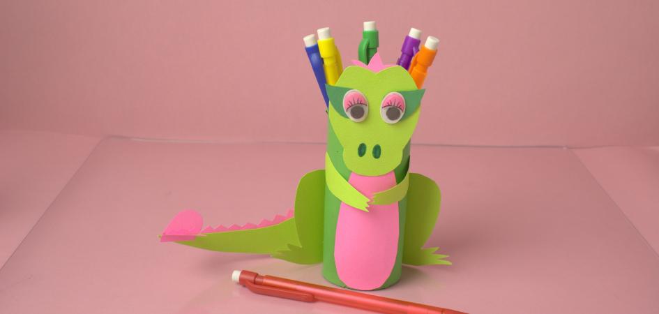 A homemade dragon pencil holder made of green paper holding colorful pencils with another pencil in front of it