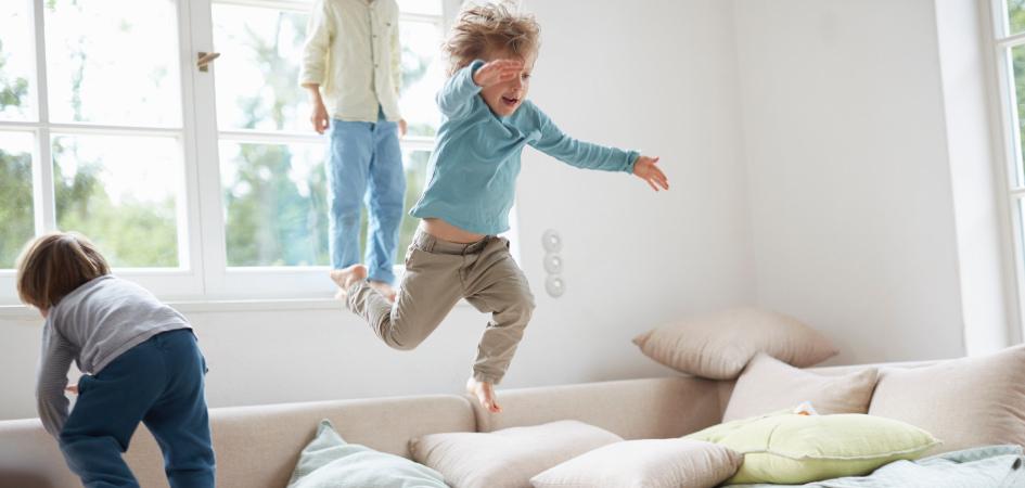 A young child jumping onto a beige sofa with cushions in the background