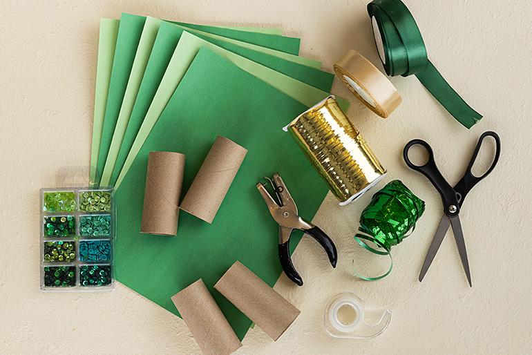 Flatlay of craft supplies for making binoculars, including green construction paper, cardboard tubes, scissors, hole punch, ribbon, sequins, and tape.