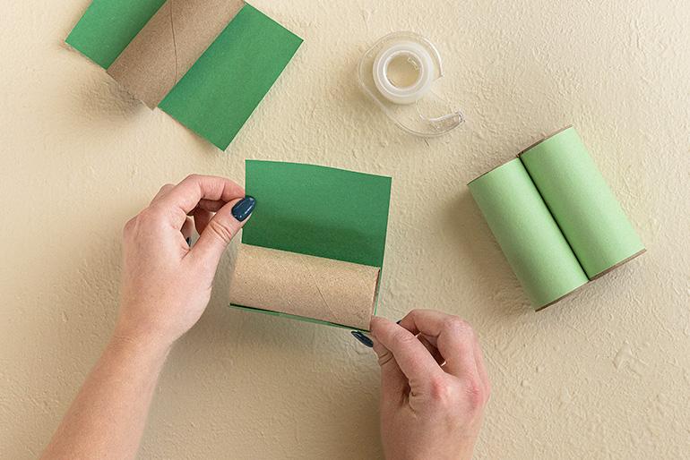 A person's hands wrapping a cardboard tube with green construction paper. Other craft supplies are visible in the background.