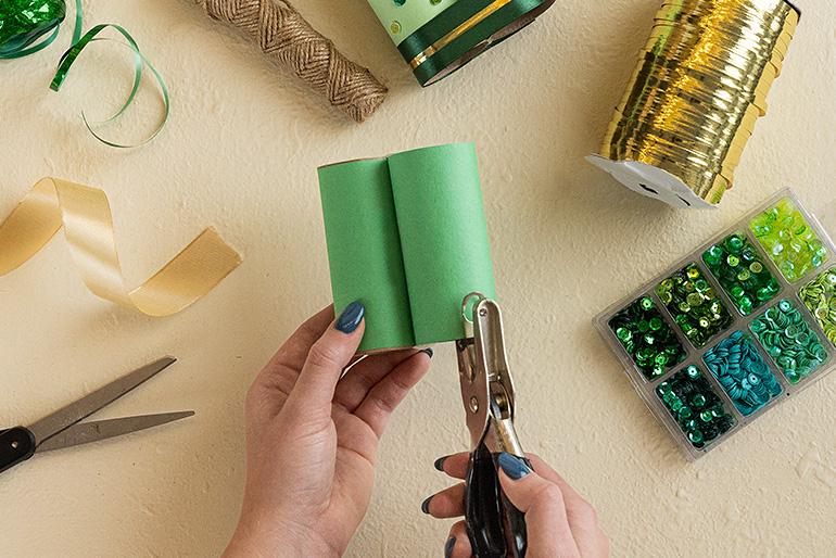 A person's hands using a hole punch on a cardboard tube wrapped in green construction paper. Other craft supplies are visible in the background.