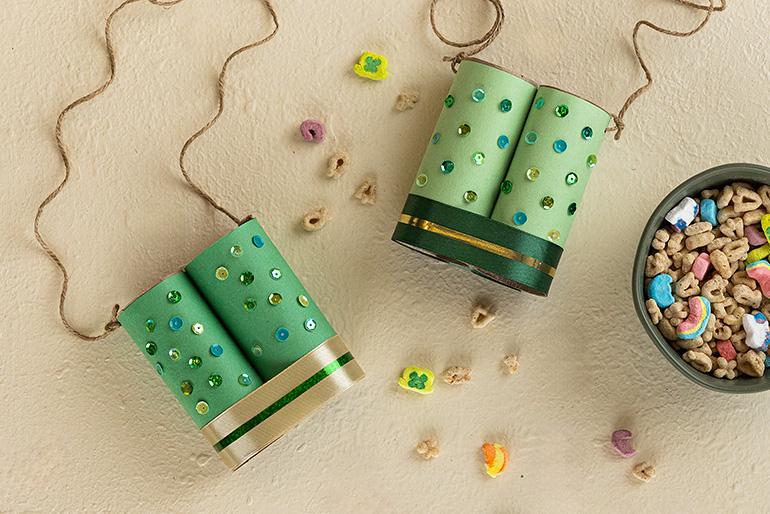 Two finished pairs of binoculars made from cardboard tubes, decorated with green paper, sequins, and ribbon. They are connected with twine. A bowl of Lucky Charms cereal is in the corner.