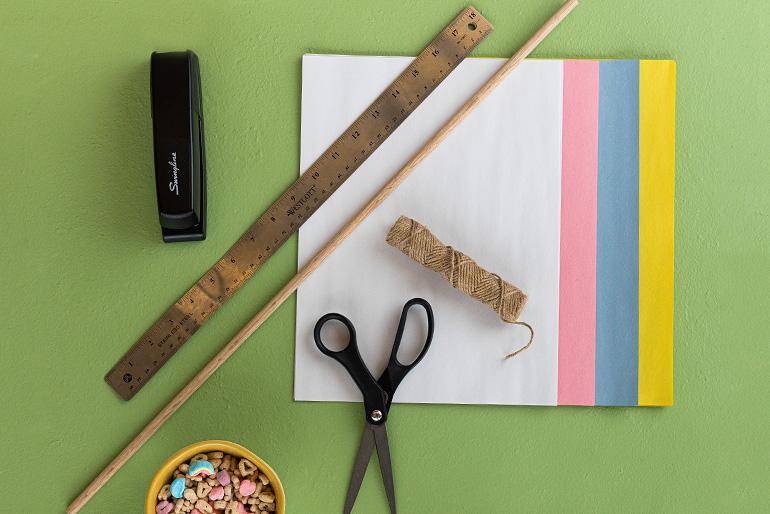 Flatlay of craft supplies on a green surface including a ruler, wooden dowel, scissors, twine, a stapler, construction paper, and a bowl of Lucky Charms cereal.