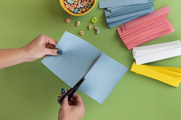 Overhead view of hands using scissors to cut a light blue piece of paper. Stacks of pink, blue, white, and yellow construction paper are nearby, along with a bowl of Lucky Charms cereal. The background is a green surface.