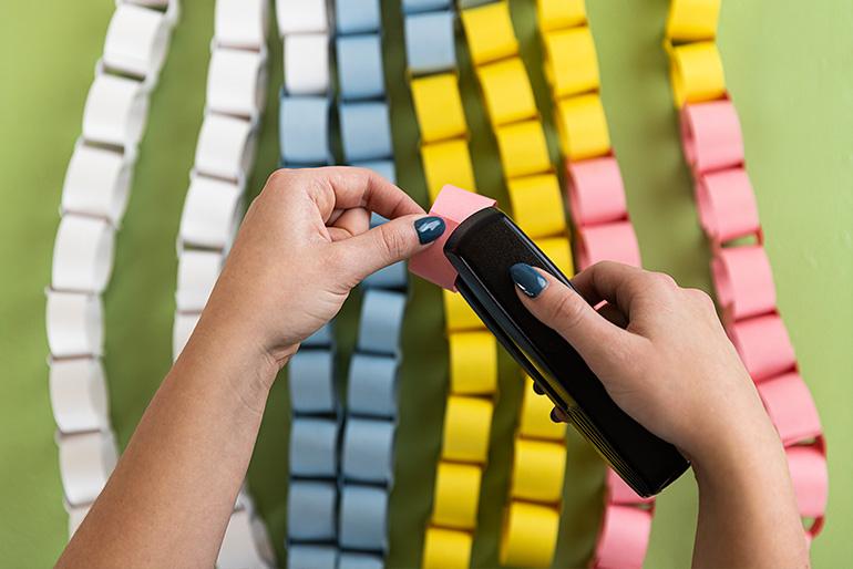 Close-up view of hands using a stapler to connect pieces of colored paper to create a paper chain. Multiple chains in pink, blue, yellow, and white are visible.