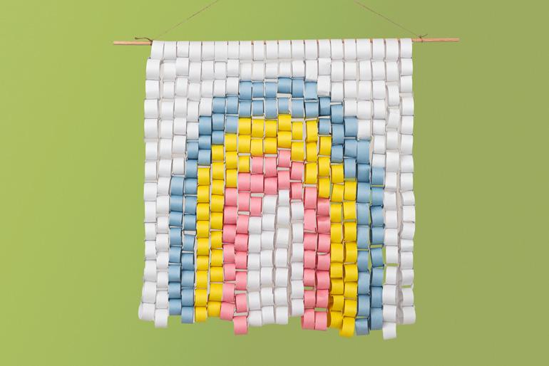 A completed rainbow-shaped wall hanging made from rolled and stapled paper chains. The wall hanging is suspended from a wooden dowel against a solid green background.