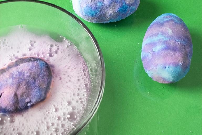 A tie-dye Lucky Charms™ Bath Bomb dissolving in a clear glass bowl of liquid.